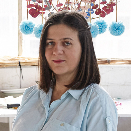 Portrait of Karolina Merska in her studio, with one of her pajaki chandelier creations hanging colourfully behind her.
