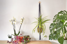 Load image into Gallery viewer, A macrame plant hanger placed next to white orchids and a fern.

